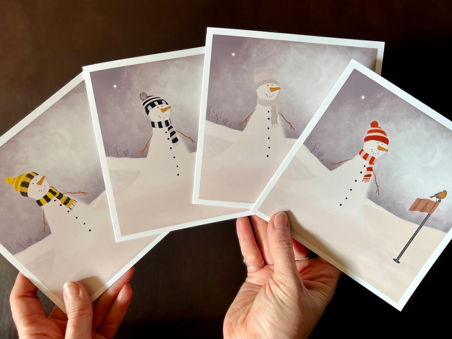 Infographic showing four artisan Christmas golf card designs featuring a snowman standing next to a golf flag on a snowy course with a robin perched on the flag. The variations show different scarf and bobble hat colours including black and amber, black and white stripes, red and white, and pale lilac and white.