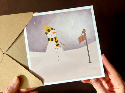 Hands holding an artisan Christmas card being removed from a kraft envelope, featuring a snowman standing next to a golf flag on a snowy golf course with a robin perched on the flag. The snowman is wearing a black and amber scarf and matching bobble hat in a sports inspired design.