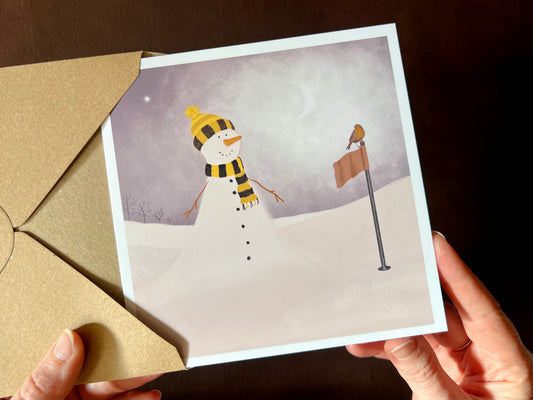 Hands holding an artisan Christmas card being removed from a kraft envelope, featuring a snowman standing next to a golf flag on a snowy golf course with a robin perched on the flag. The snowman is wearing a black and amber scarf and matching bobble hat in a sports inspired design.