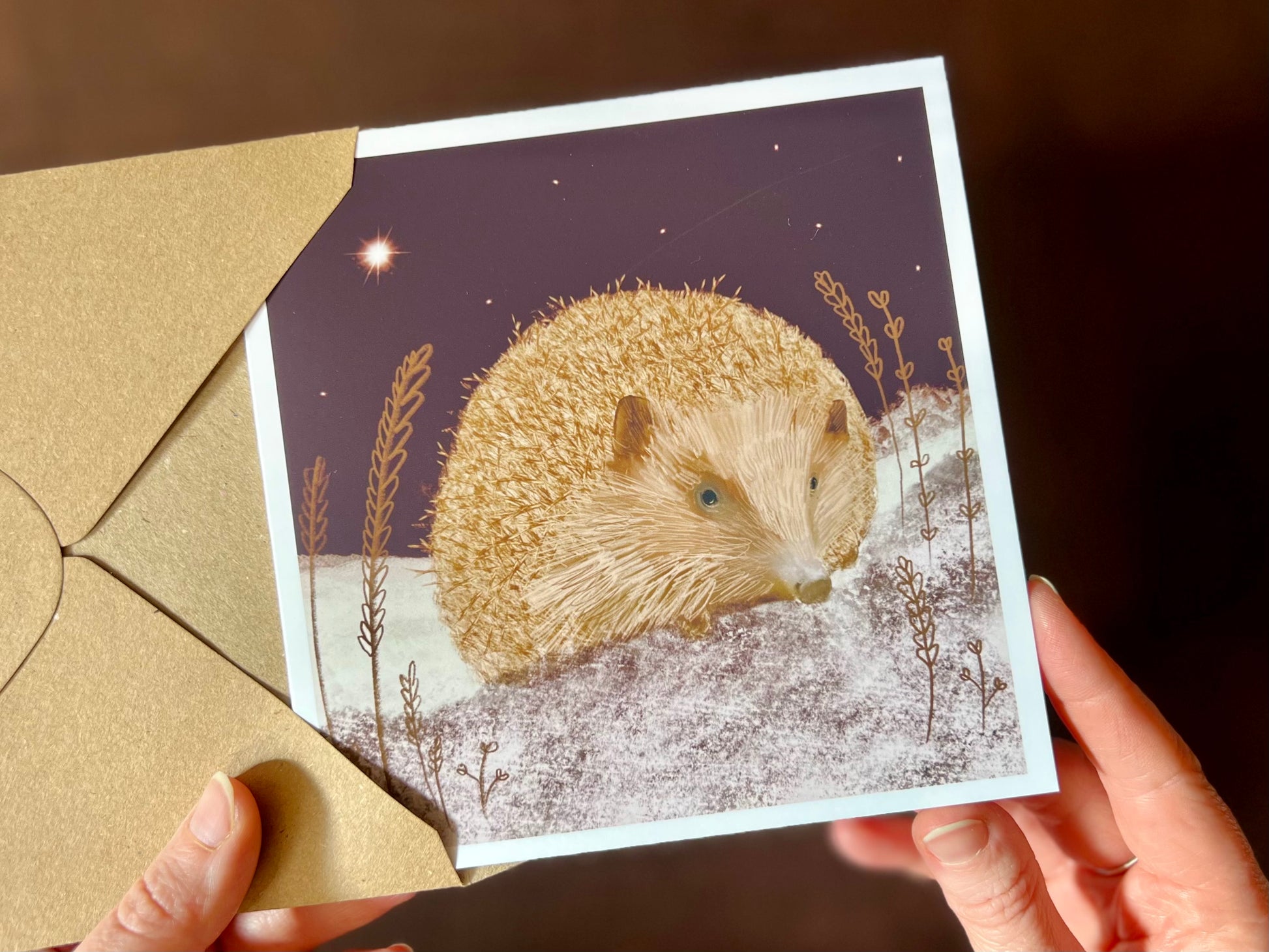 Hands holding an artisan Christmas card being removed from a kraft envelope, featuring a magical illustration of a hedgehog sitting on lightly snow-covered ground beneath a dark purple night sky with a shining star. The design has a festive winter atmosphere.