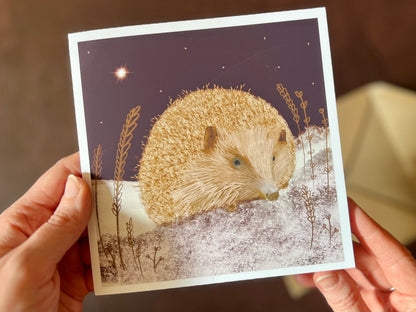 Close up image of hands holding an artisan Christmas card being removed from a kraft envelope, featuring a magical hedgehog illustration sitting on lightly snow-covered ground beneath a dark purple night sky with a shining star.