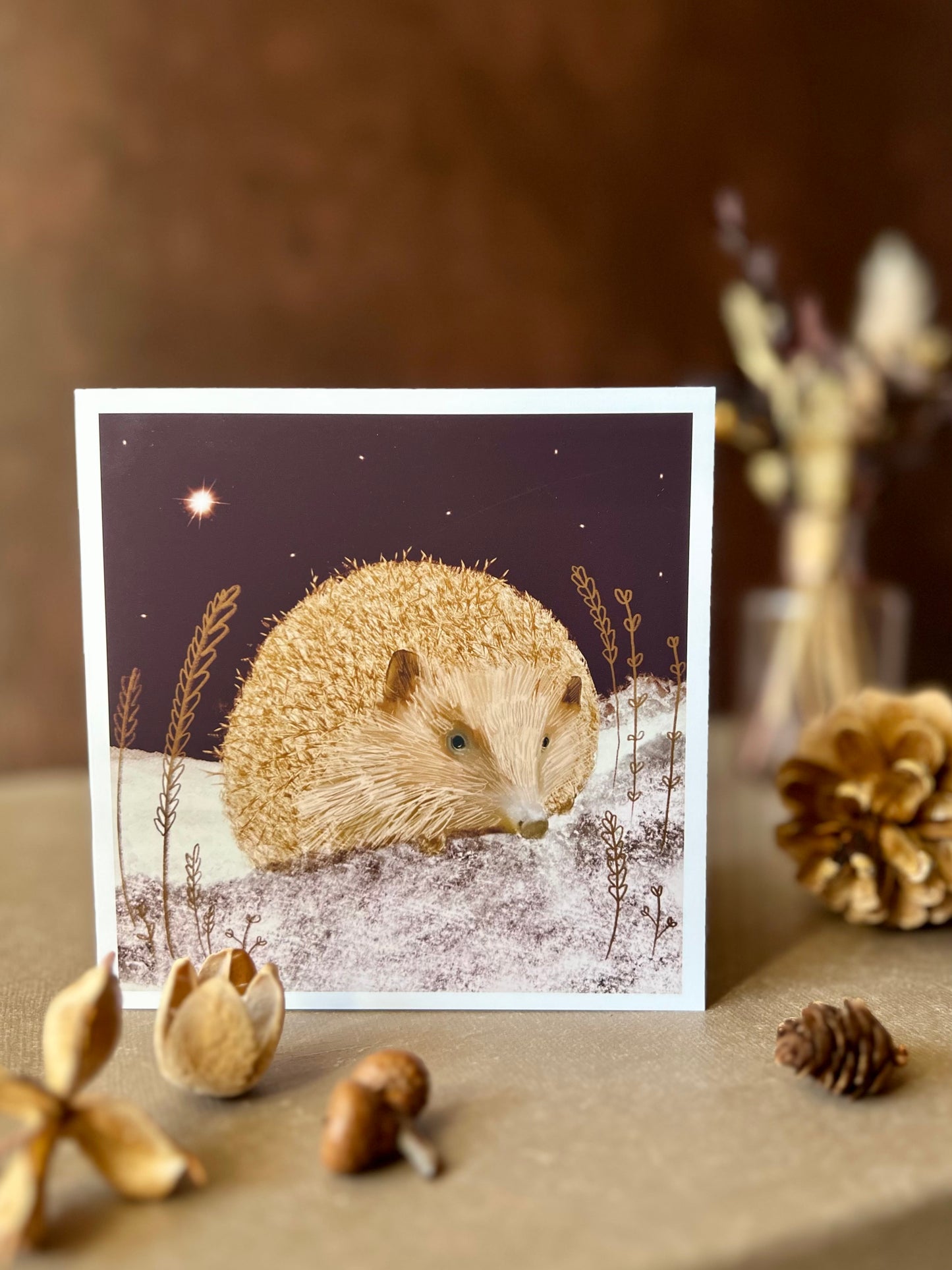 Artisan Christmas card featuring a magical hedgehog illustration sitting on lightly snow-covered ground beneath a dark purple night sky with a shining star, shown standing upright on a tabletop. The foreground includes woodland pine cones and acorns, with dried flowers softly blurred in the background.