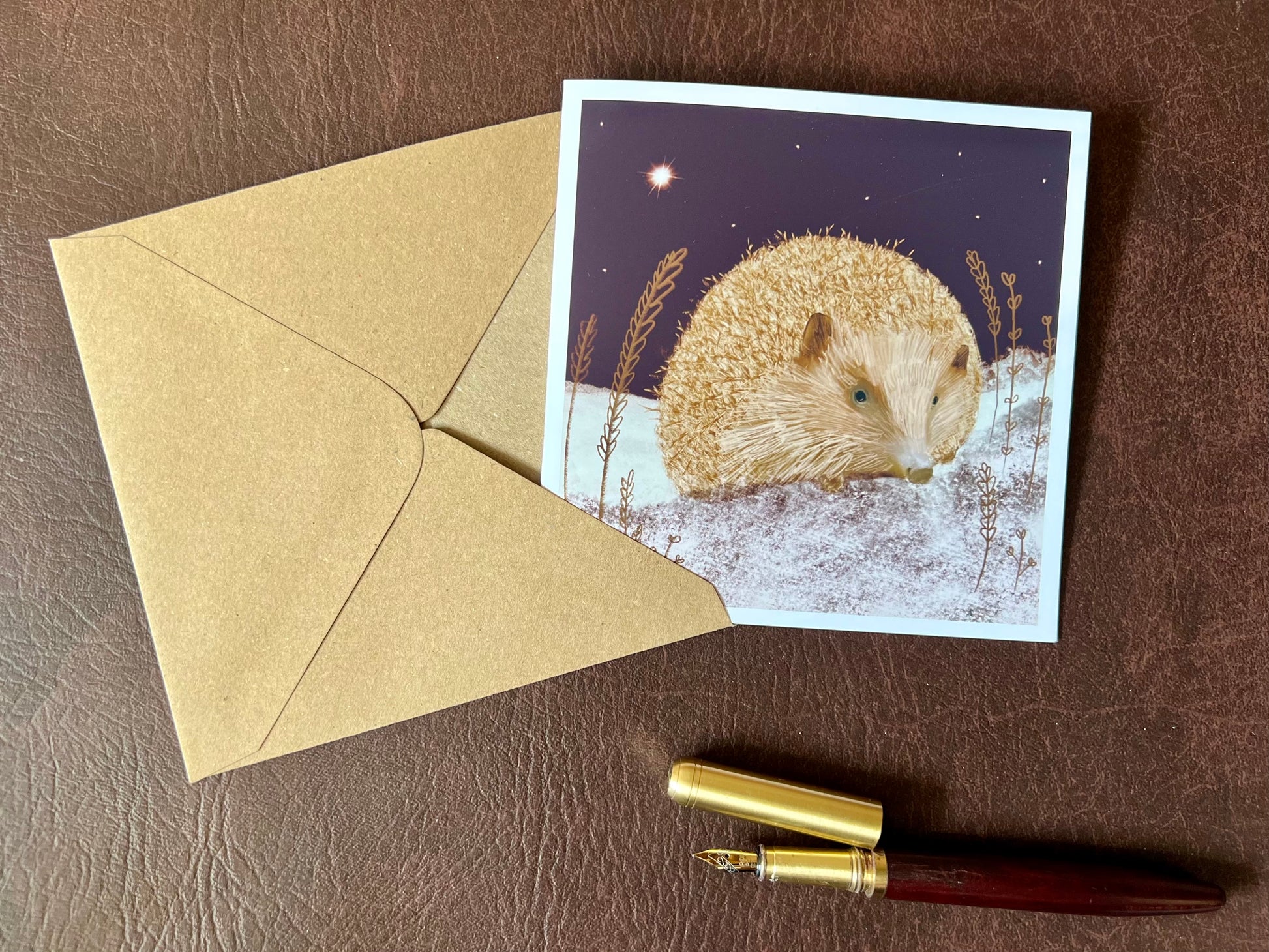 Flat lay image of an artisan Christmas card featuring a magical hedgehog illustration sitting on lightly snow-covered ground beneath a dark purple night sky with a shining star, shown laid flat on a tabletop with its kraft envelope.