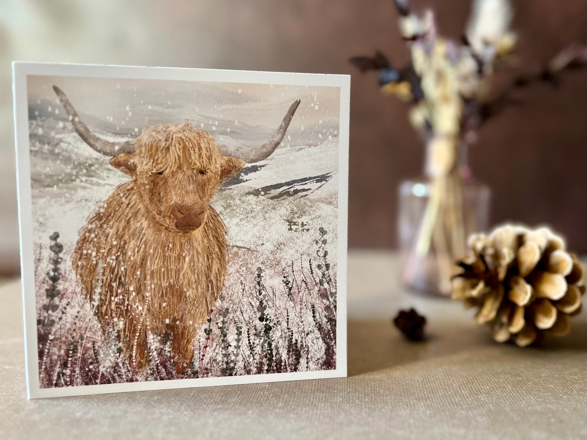 Artisan Christmas card featuring a single highland cow standing in a snowy heather moorland landscape, shown standing upright on a tabletop in a styled seasonal display with no text on the front.