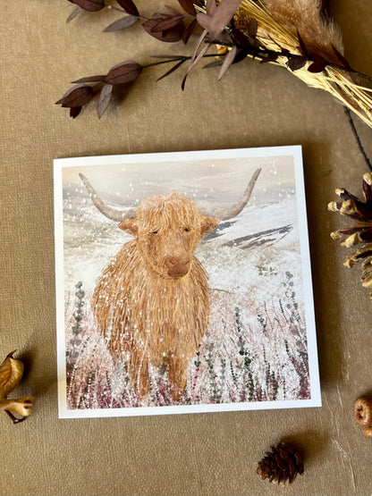 Flat lay image of an artisan Christmas card featuring a single highland cow standing in a snowy heather moorland landscape with no text, shown laid flat on a tabletop surrounded by dried flowers and pine cones in a styled winter composition.