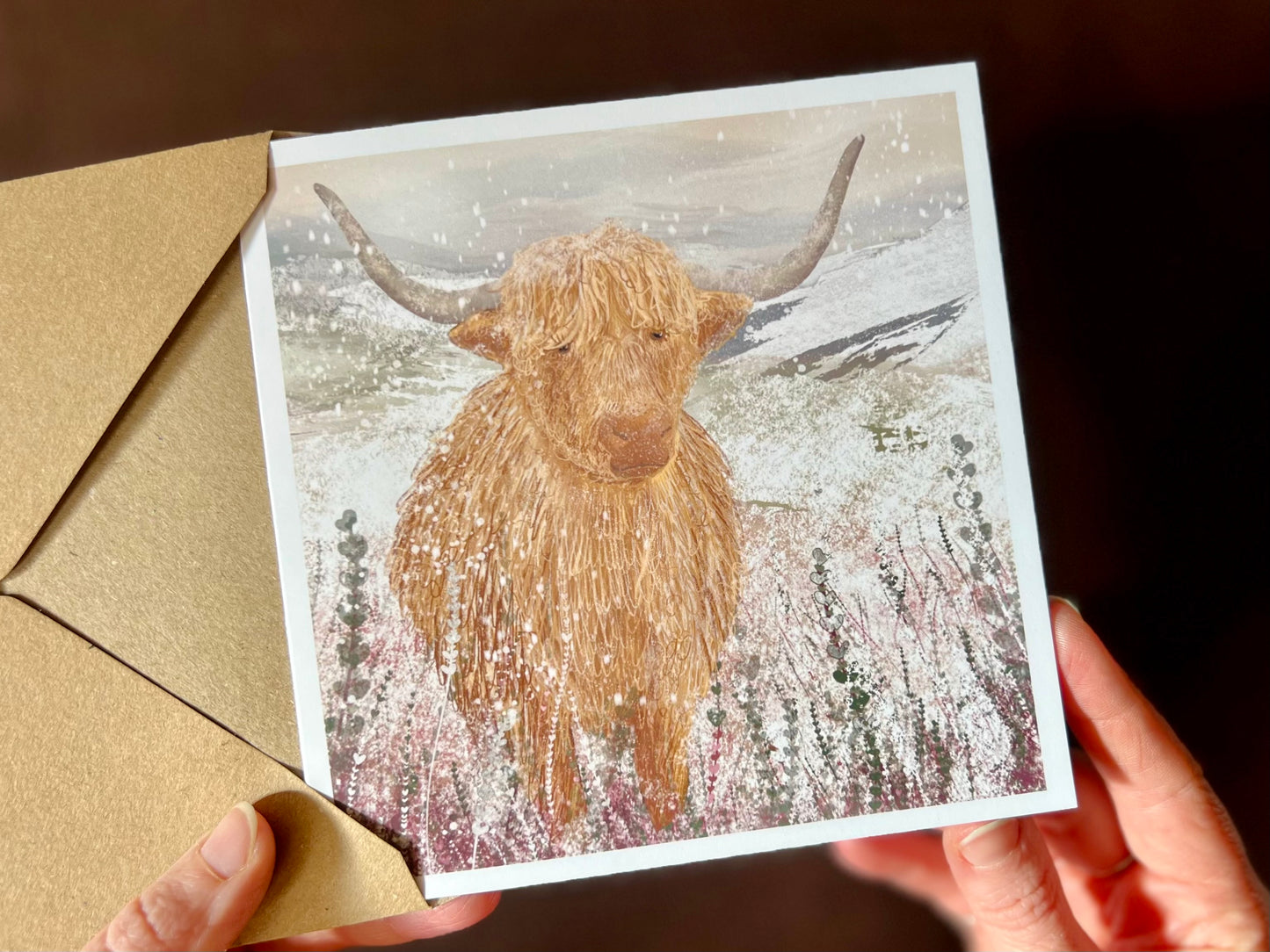 Hands holding an artisan Christmas card being removed from a kraft envelope, featuring a single highland cow standing in a snowy heather moorland landscape with a winter atmosphere. The design is a festive seasonal variation of the original illustration with no text on the front.