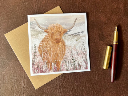 Flat lay image of an artisan Christmas card featuring a single highland cow standing in a snowy heather moorland landscape with no text, shown laid flat on a leatherette tabletop in a styled seasonal composition.