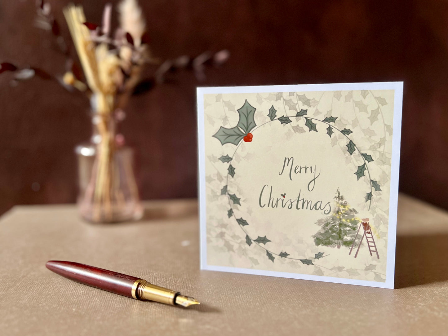 Artisan Christmas card featuring a creamy pale yellow background with a holly wreath border and handwritten style 'Merry Christmas' text, with a small mouse on a stepladder decorating a Christmas tree in the corner, shown standing upright on a tabletop in a styled display.