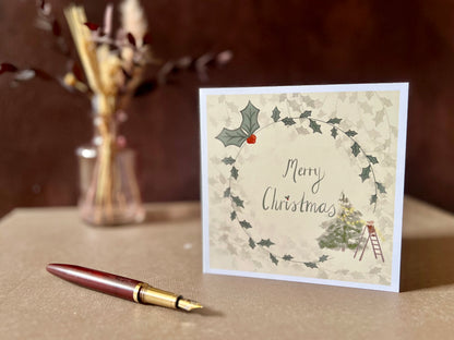 Artisan Christmas card featuring a creamy pale yellow background with a holly wreath border and handwritten style 'Merry Christmas' text, with a small mouse on a stepladder decorating a Christmas tree in the corner, shown standing upright on a tabletop in a styled display.