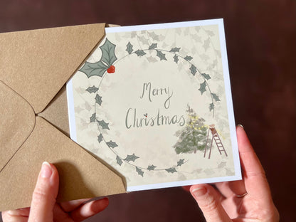 Hands holding an artisan Christmas card being removed from a kraft envelope, featuring a creamy pale yellow background with a holly wreath border and handwritten style 'Merry Christmas' text. In the corner is an illustration of a mouse on a stepladder decorating a forest Christmas tree.