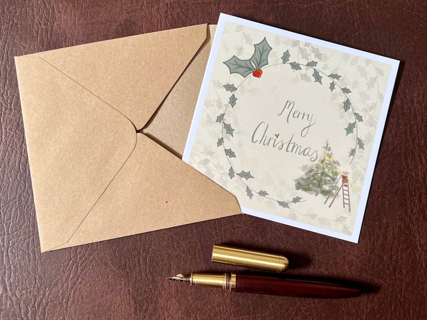 Flat lay image of an artisan Christmas card featuring a creamy pale yellow background with a holly wreath border and handwritten style 'Merry Christmas' text, with a small mouse on a stepladder decorating a Christmas tree in the corner, shown laid flat on a tabletop.