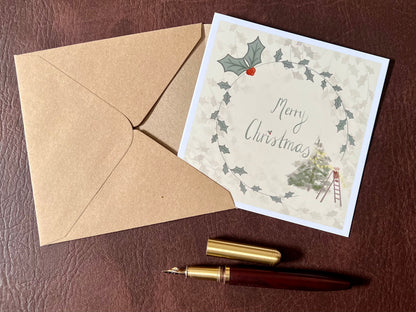 Flat lay image of an artisan Christmas card featuring a creamy pale yellow background with a holly wreath border and handwritten style 'Merry Christmas' text, with a small mouse on a stepladder decorating a Christmas tree in the corner, shown laid flat on a tabletop.