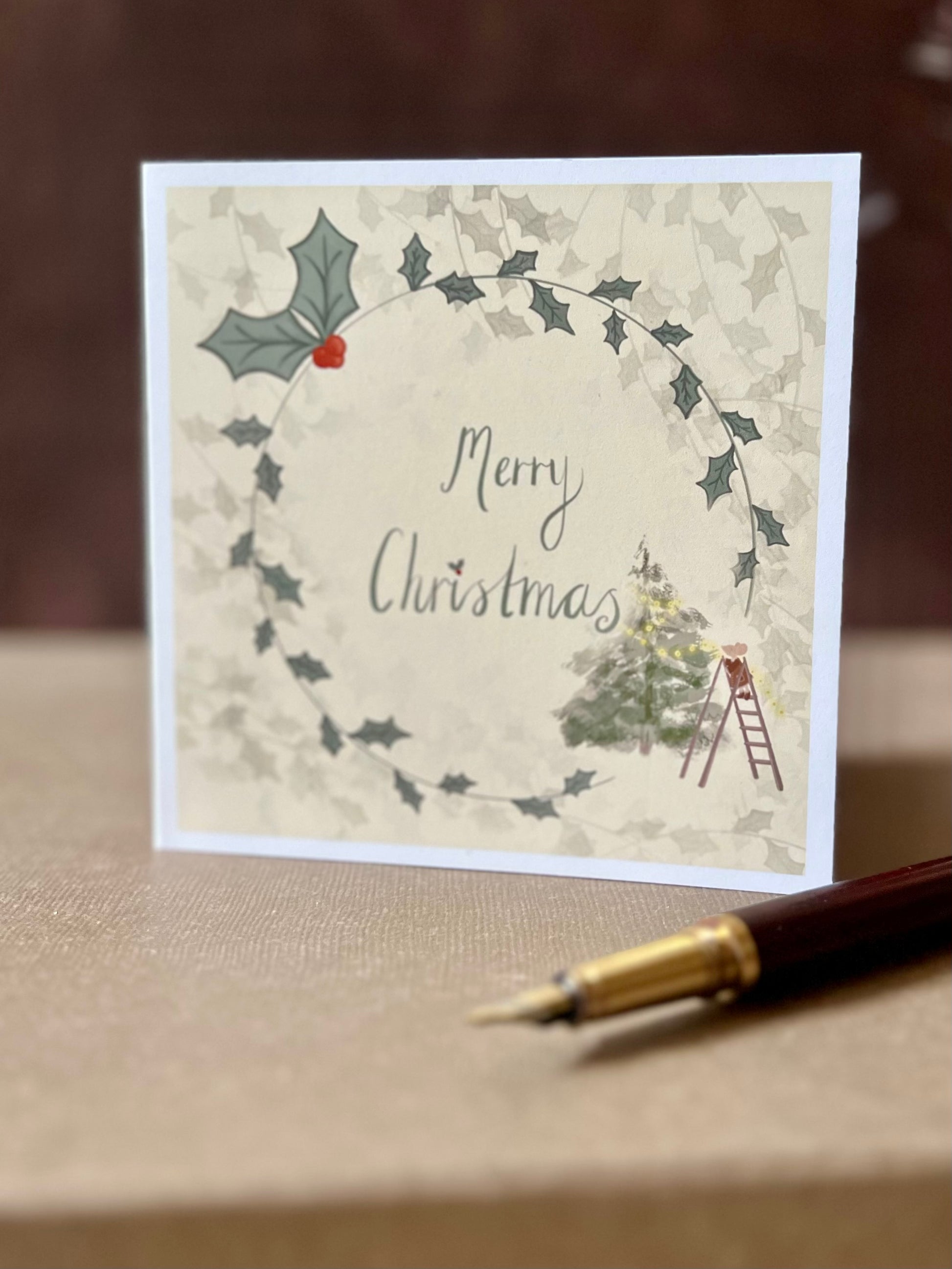 Portrait oriented artisan Christmas card featuring a creamy pale yellow background with a holly wreath border and handwritten style 'Merry Christmas' text, with a small mouse on a stepladder decorating a Christmas tree in the corner, shown standing upright in a styled tabletop display.