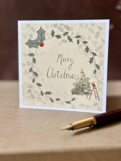 Portrait oriented artisan Christmas card featuring a creamy pale yellow background with a holly wreath border and handwritten style 'Merry Christmas' text, with a small mouse on a stepladder decorating a Christmas tree in the corner, shown standing upright in a styled tabletop display.