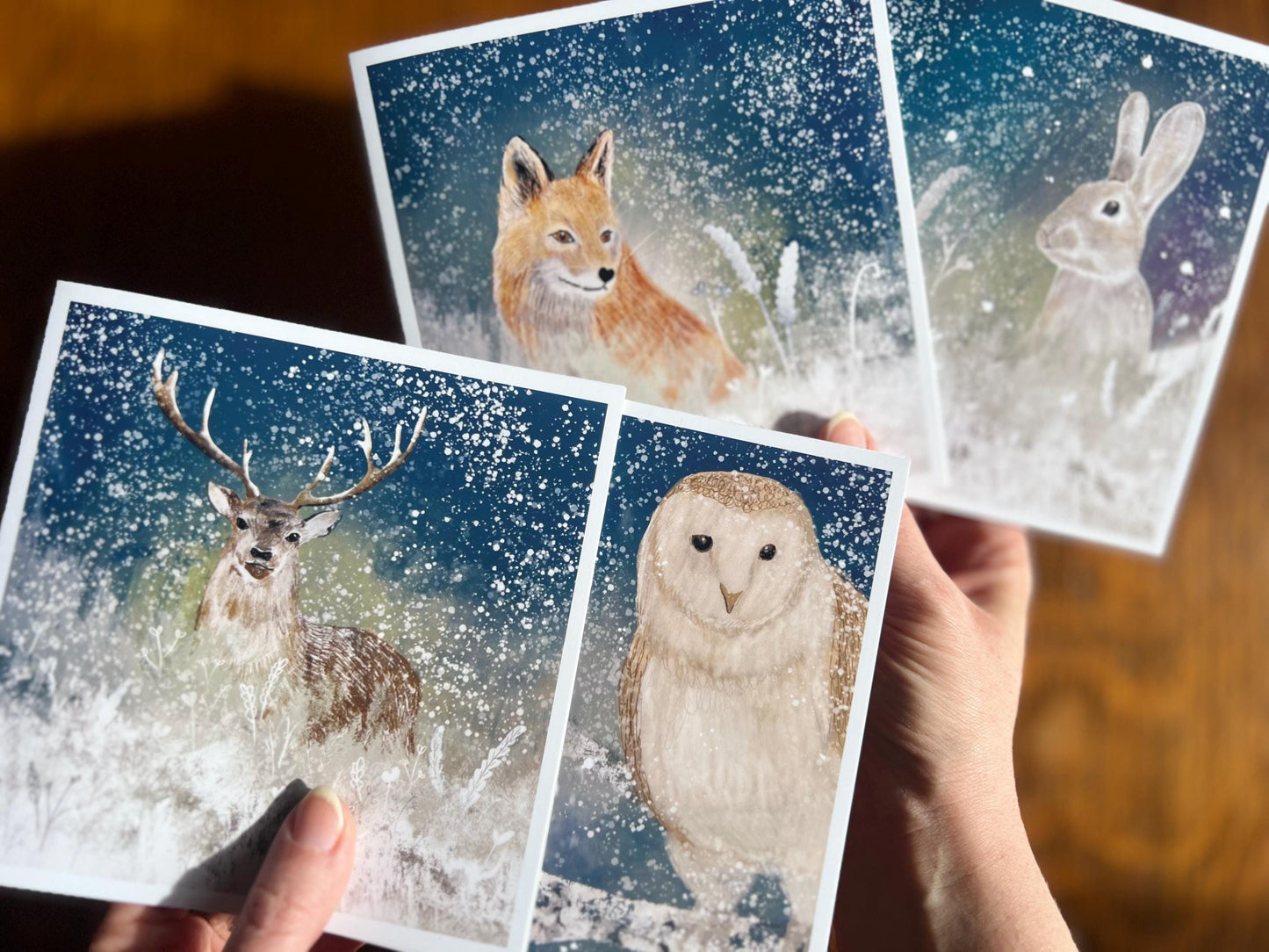 Hands holding a selection of four artisan Christmas cards featuring nighttime winter wildlife designs, shown in a fan-shaped display to highlight the collection of fox, reindeer, deer, and rabbit under snowy night skies.