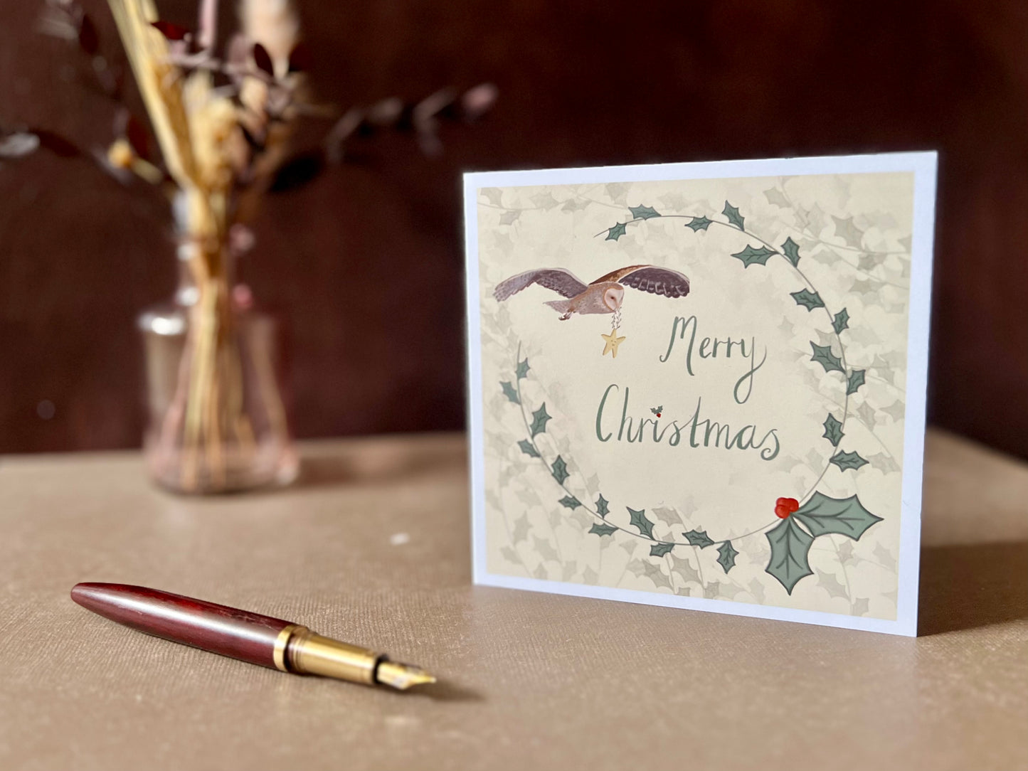 Artisan Christmas card featuring a creamy pale yellow background with a holly wreath border and handwritten style 'Merry Christmas' text, with an illustration of an owl flying through the sky carrying a smiling mouse, shown standing upright on a tabletop in a styled display.