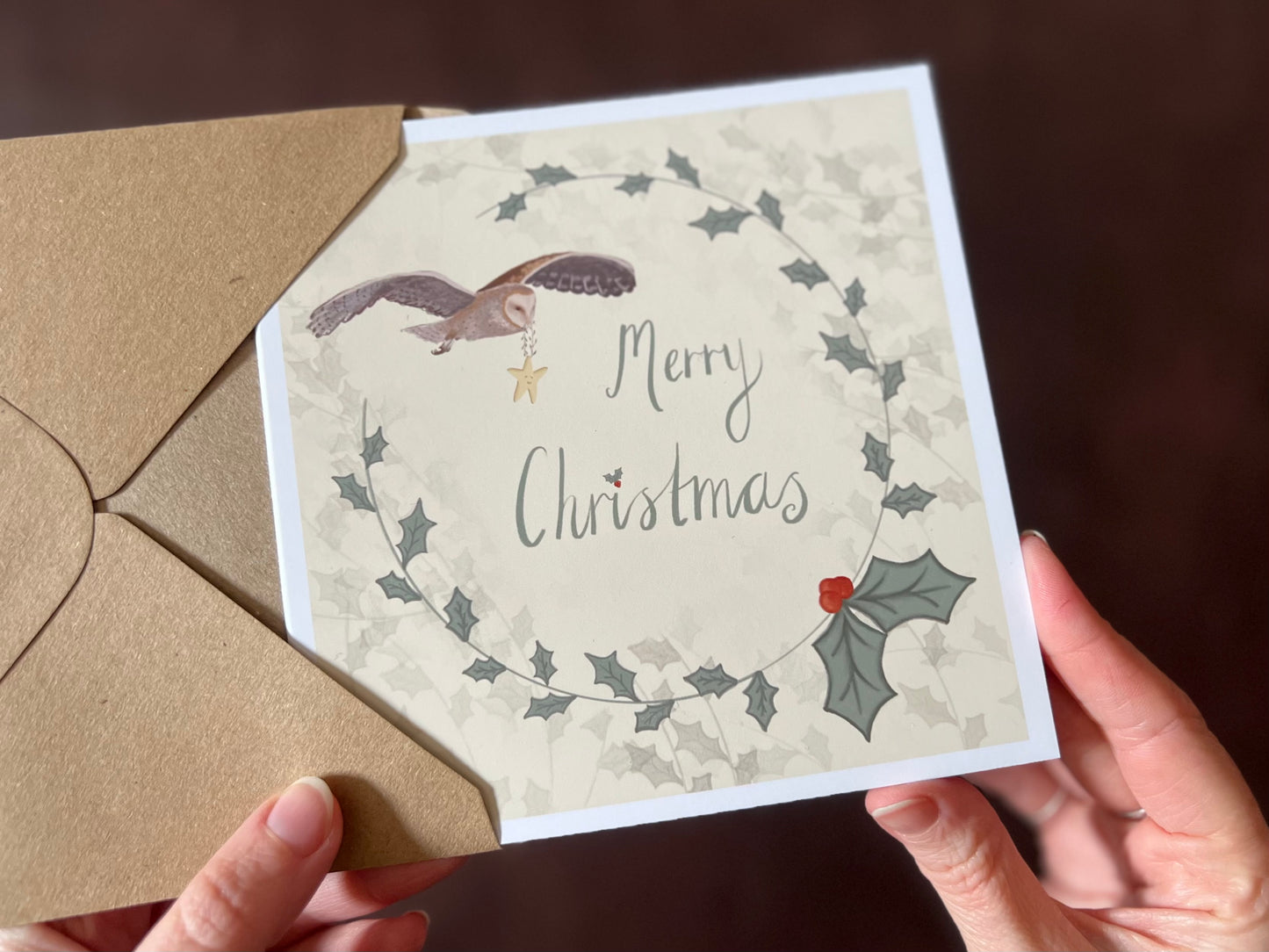 Hands holding an artisan Christmas card being removed from a kraft envelope, featuring a creamy pale yellow background with a holly wreath border and handwritten style 'Merry Christmas' text. In the top corner, an owl is shown flying through the sky carrying a smiling mouse.