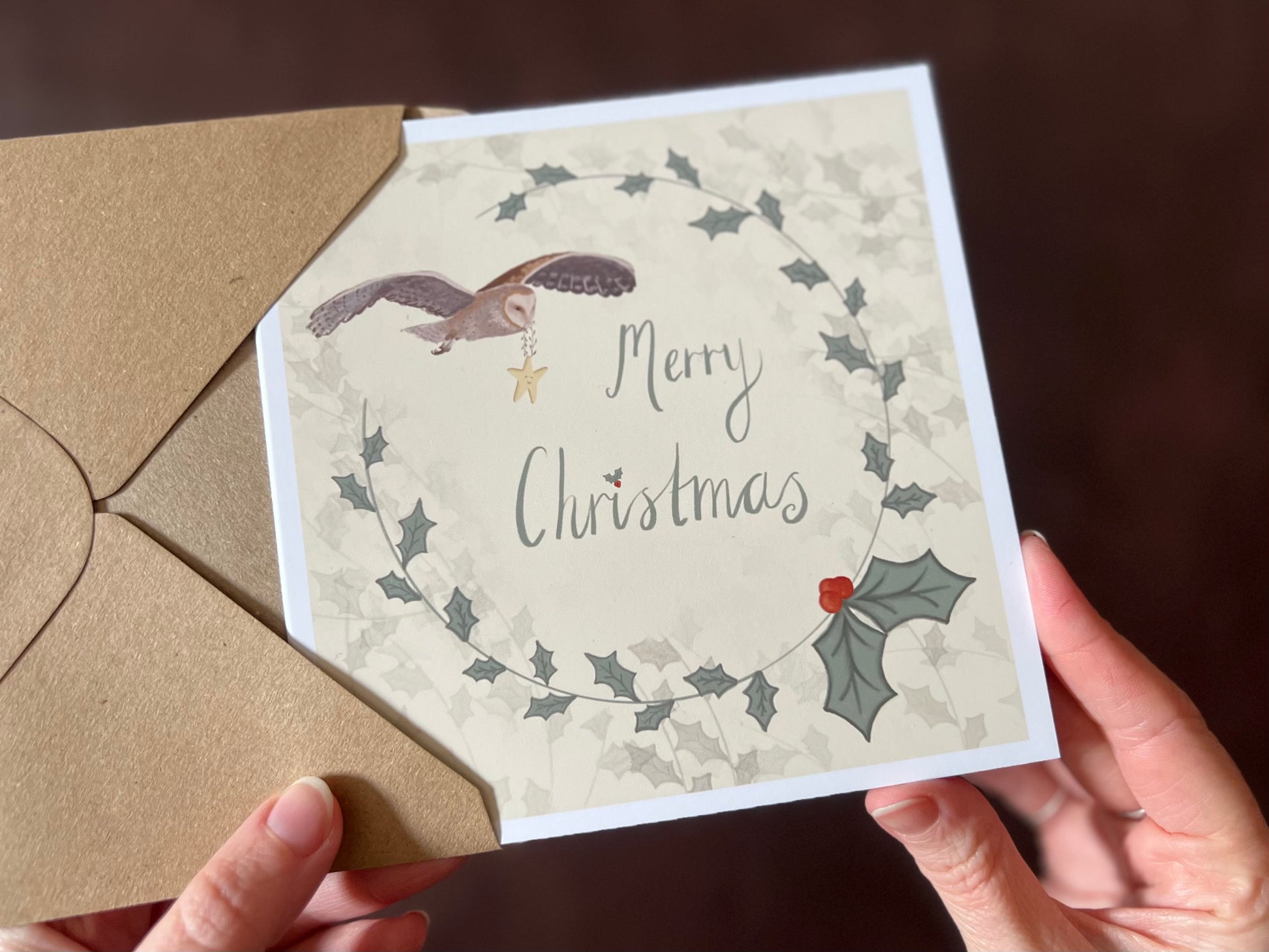 Hands holding an artisan Christmas card being removed from a kraft envelope, featuring a creamy pale yellow background with a holly wreath border and handwritten style 'Merry Christmas' text. In the top corner, an owl is shown flying through the sky carrying a smiling mouse.