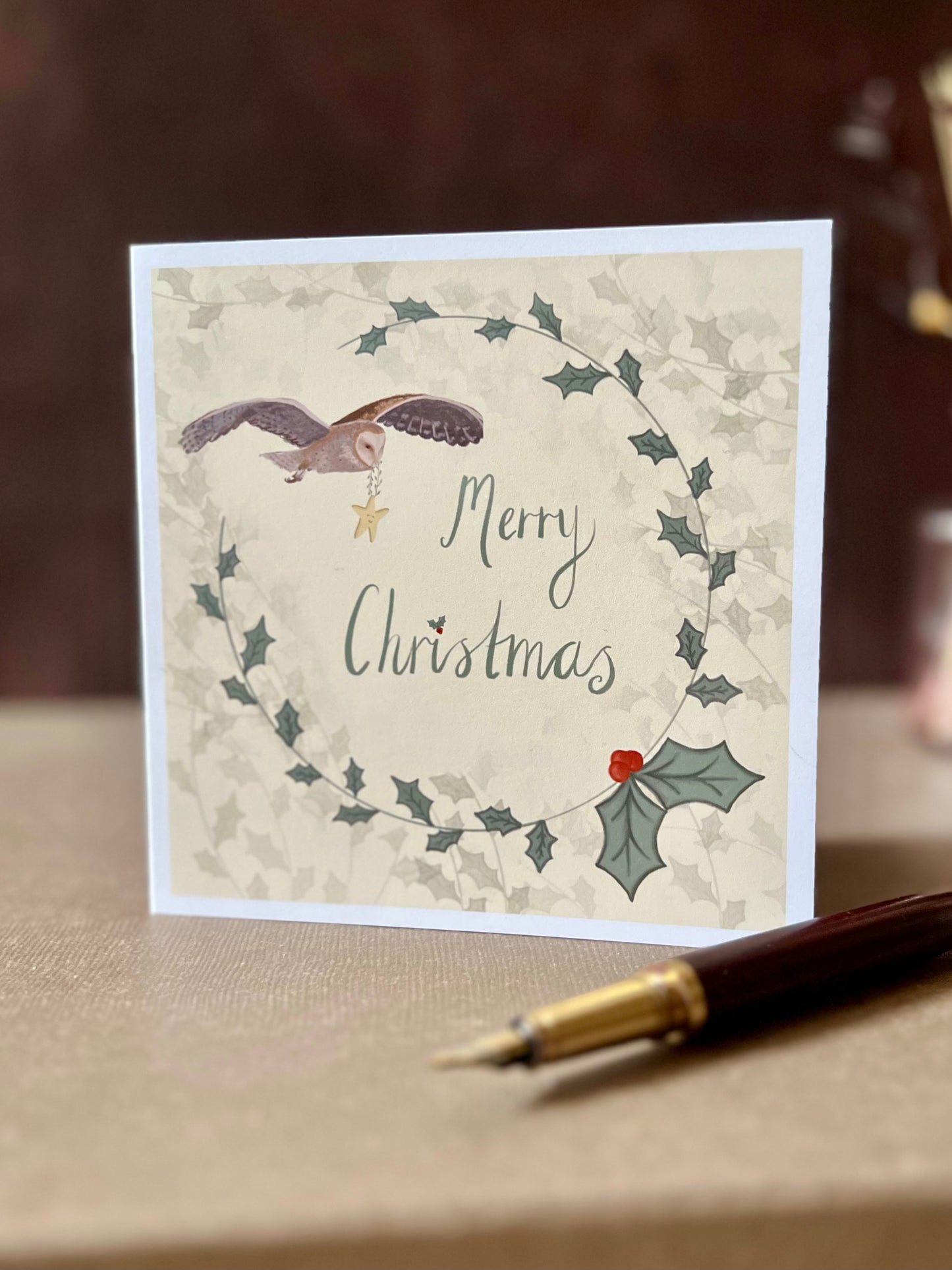 Portrait oriented artisan Christmas card featuring a creamy pale yellow background with a holly wreath border and handwritten style 'Merry Christmas' text, with an illustration of an owl flying through the sky carrying a smiling mouse, shown standing upright in a styled tabletop display.