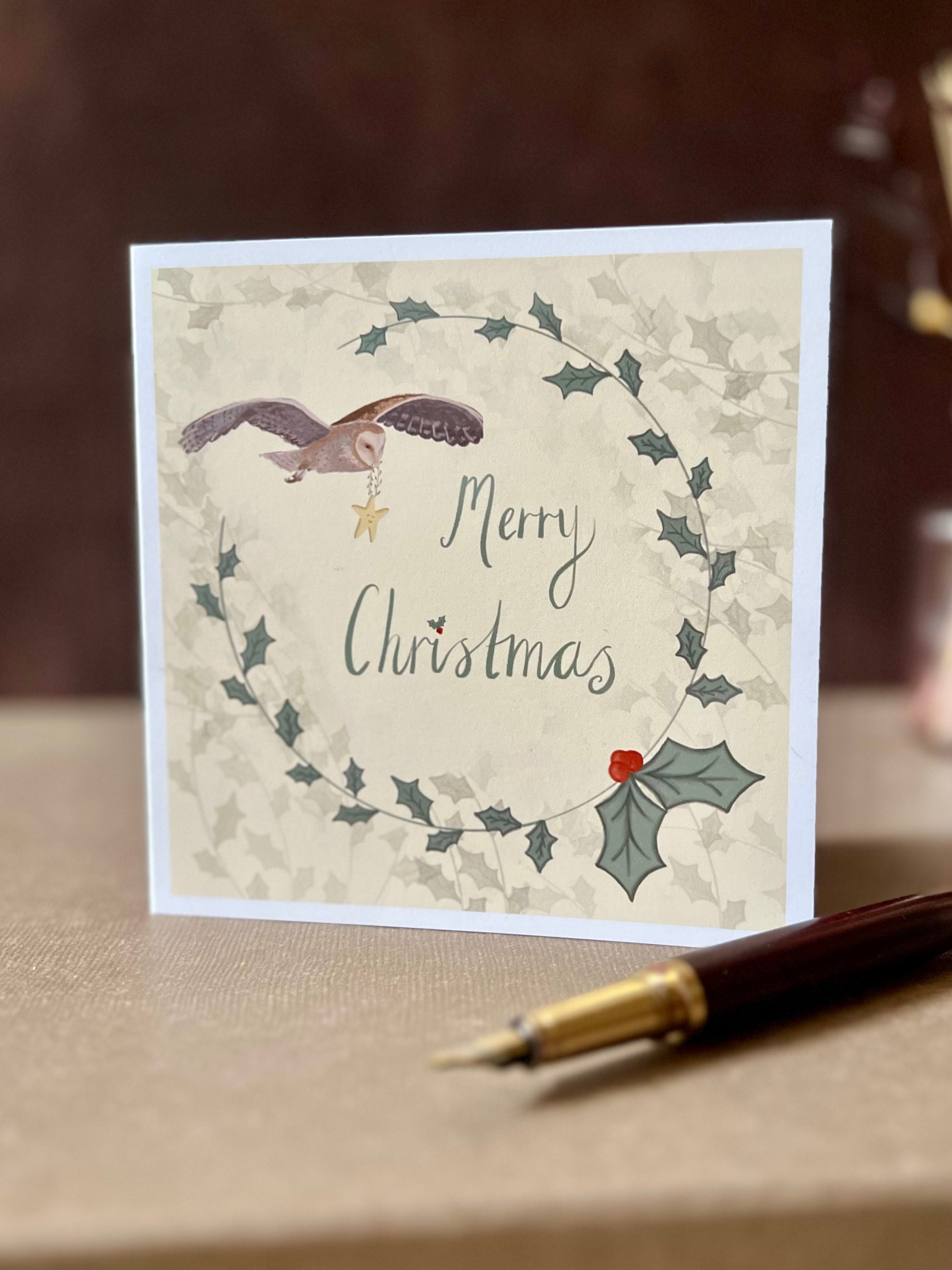 Portrait oriented artisan Christmas card featuring a creamy pale yellow background with a holly wreath border and handwritten style 'Merry Christmas' text, with an illustration of an owl flying through the sky carrying a smiling mouse, shown standing upright in a styled tabletop display.