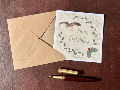 Flat lay image of an artisan Christmas card featuring a creamy pale yellow background with a holly wreath border and handwritten style 'Merry Christmas' text, with an illustration of an owl flying through the sky carrying a smiling mouse, shown laid flat on a tabletop in a styled composition.