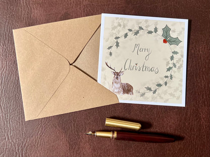 Flat lay image of an artisan Christmas card featuring a creamy pale yellow background with a holly wreath border and handwritten style 'Merry Christmas' text, with a reindeer illustration in the bottom left corner, shown laid flat on a tabletop in a styled festive composition.