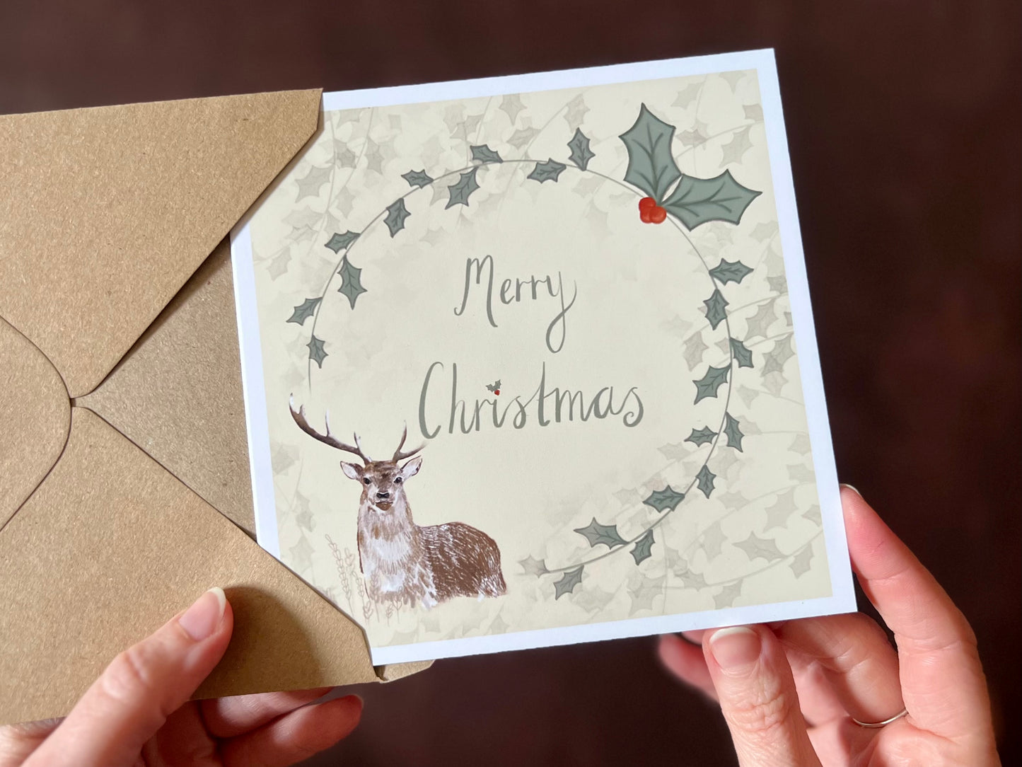 Hands holding an artisan Christmas card being removed from a kraft envelope, featuring a creamy pale yellow background with a holly wreath border and handwritten style 'Merry Christmas' text. A reindeer illustration appears in the bottom left corner, creating a traditional festive design.