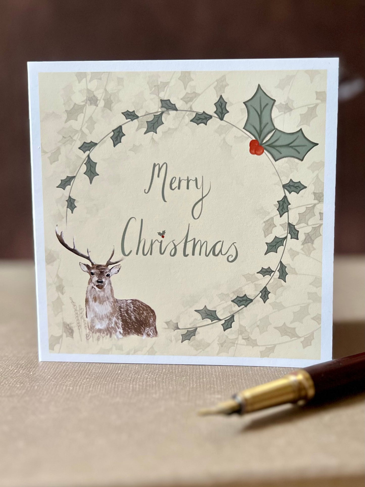 Portrait oriented artisan Christmas card featuring a creamy pale yellow background with a holly wreath border and handwritten style 'Merry Christmas' text, with a reindeer illustration in the bottom left corner, shown standing upright in a styled tabletop display.