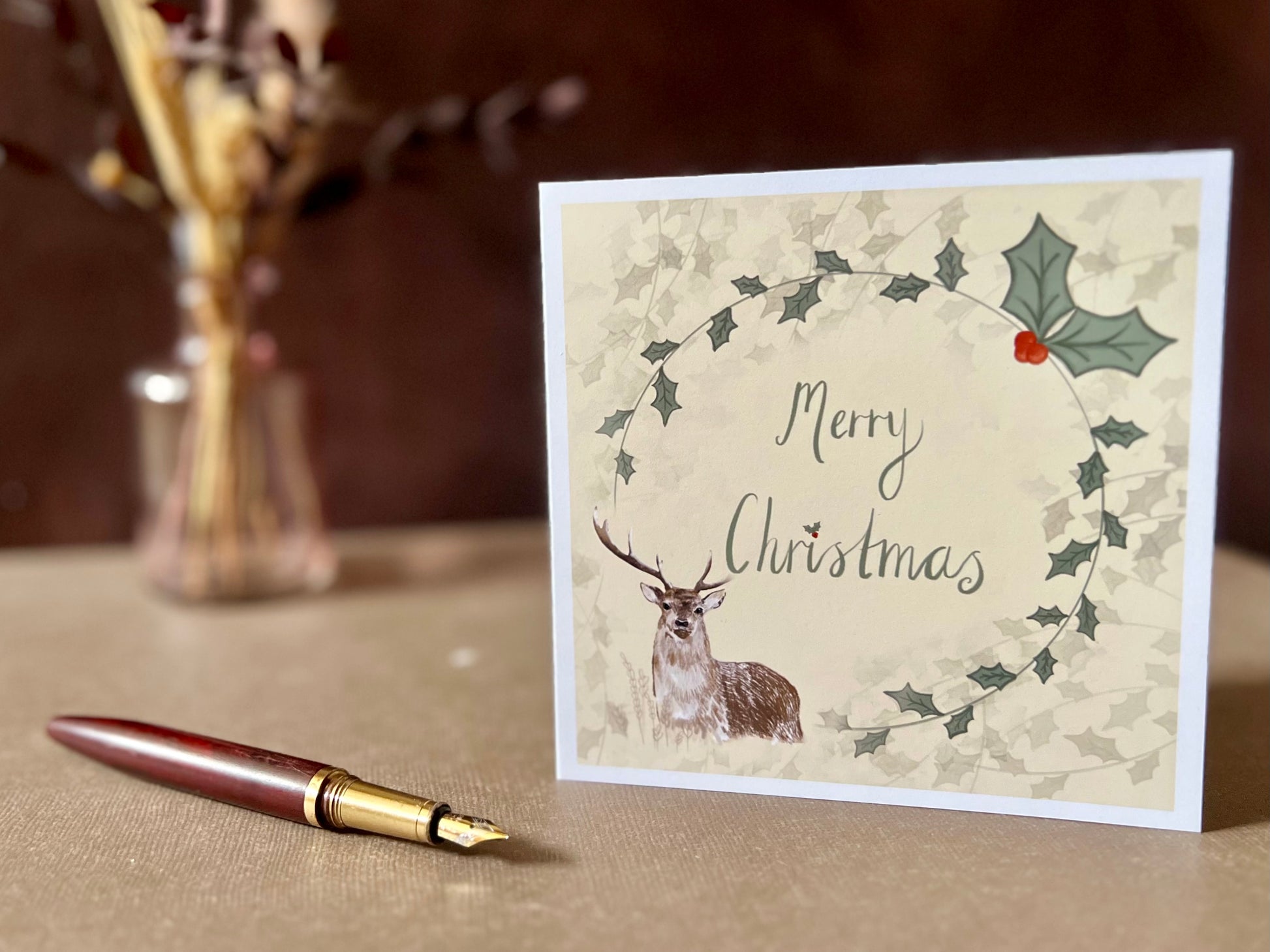 Artisan Christmas card featuring a creamy pale yellow background with a holly wreath border and handwritten style 'Merry Christmas' text, with a reindeer illustration in the bottom left corner, shown standing upright on a tabletop in a styled festive display.