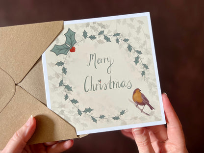 Close up image of an artisan Christmas card featuring a creamy pale yellow background with a holly wreath border and handwritten style 'Merry Christmas' text, with a robin illustration in the bottom left corner.