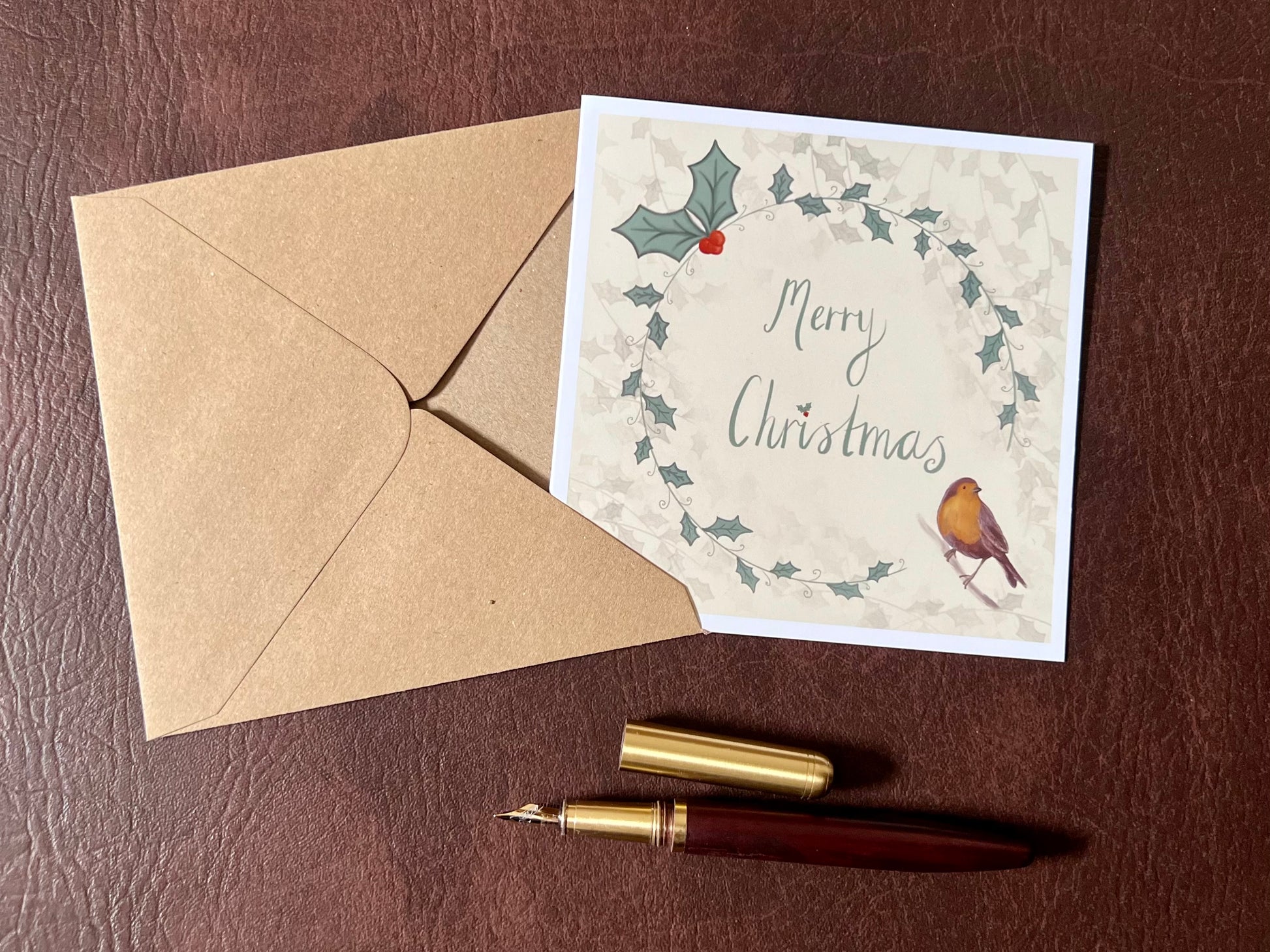 Flat lay image of an artisan Christmas card featuring a creamy pale yellow background with a holly wreath border and handwritten style 'Merry Christmas' text, with a robin illustration in the bottom left corner, shown laid flat on a tabletop in a styled festive composition.