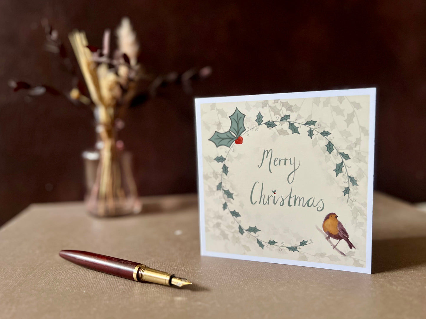 Artisan Christmas card featuring a creamy pale yellow background with a holly wreath border and handwritten style 'Merry Christmas' text, with a robin illustration in the bottom left corner, shown standing upright on a tabletop in a styled festive display.