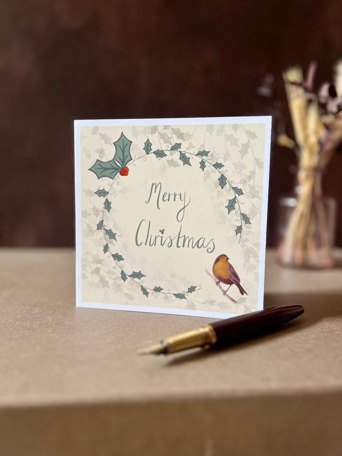 Portrait oriented artisan Christmas card featuring a creamy pale yellow background with a holly wreath border and handwritten style 'Merry Christmas' text, with a robin illustration in the bottom left corner, shown standing upright on a tabletop in a styled festive display.