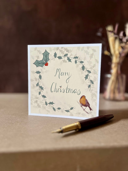Portrait oriented artisan Christmas card featuring a creamy pale yellow background with a holly wreath border and handwritten style 'Merry Christmas' text, with a robin illustration in the bottom left corner, shown standing upright on a tabletop in a styled festive display.