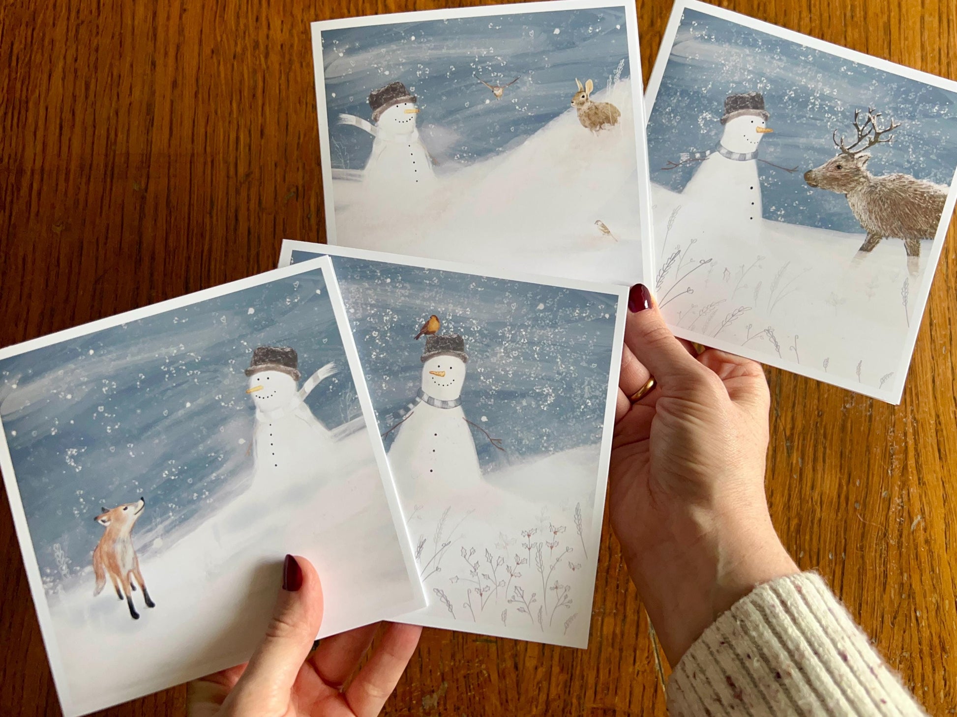 Hands holding a selection of four artisan Christmas cards featuring snowman-themed designs, shown in a fan-shaped display to highlight the festive winter collection.