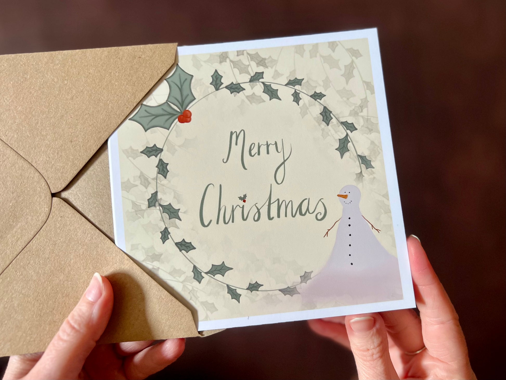 Close up image of an artisan Christmas card featuring a creamy pale yellow background with a holly wreath border and handwritten style 'Merry Christmas' text, with a snowman illustration in the corner.
