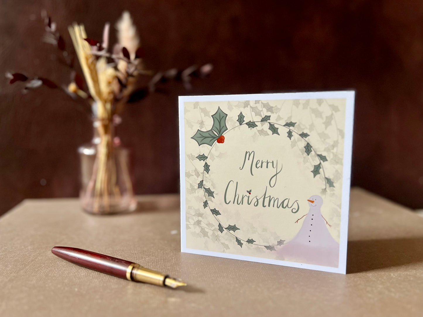 Artisan Christmas card featuring a creamy pale yellow background with a holly wreath border and handwritten style 'Merry Christmas' text, with a snowman illustration in the corner, shown standing upright on a tabletop in a styled festive display.
