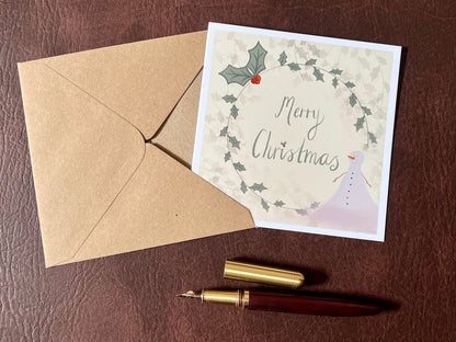 Flat lay image of an artisan Christmas card featuring a creamy pale yellow background with a holly wreath border and handwritten style 'Merry Christmas' text, with a snowman illustration in the corner, shown laid flat on a tabletop in a styled festive composition.