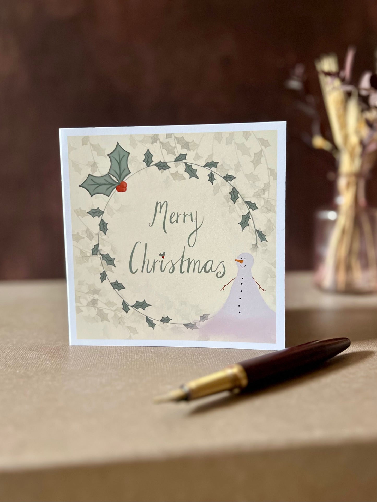 Portrait oriented artisan Christmas card featuring a creamy pale yellow background with a holly wreath border and handwritten style 'Merry Christmas' text, with a snowman illustration in the corner, shown standing upright on a tabletop in a styled festive display.