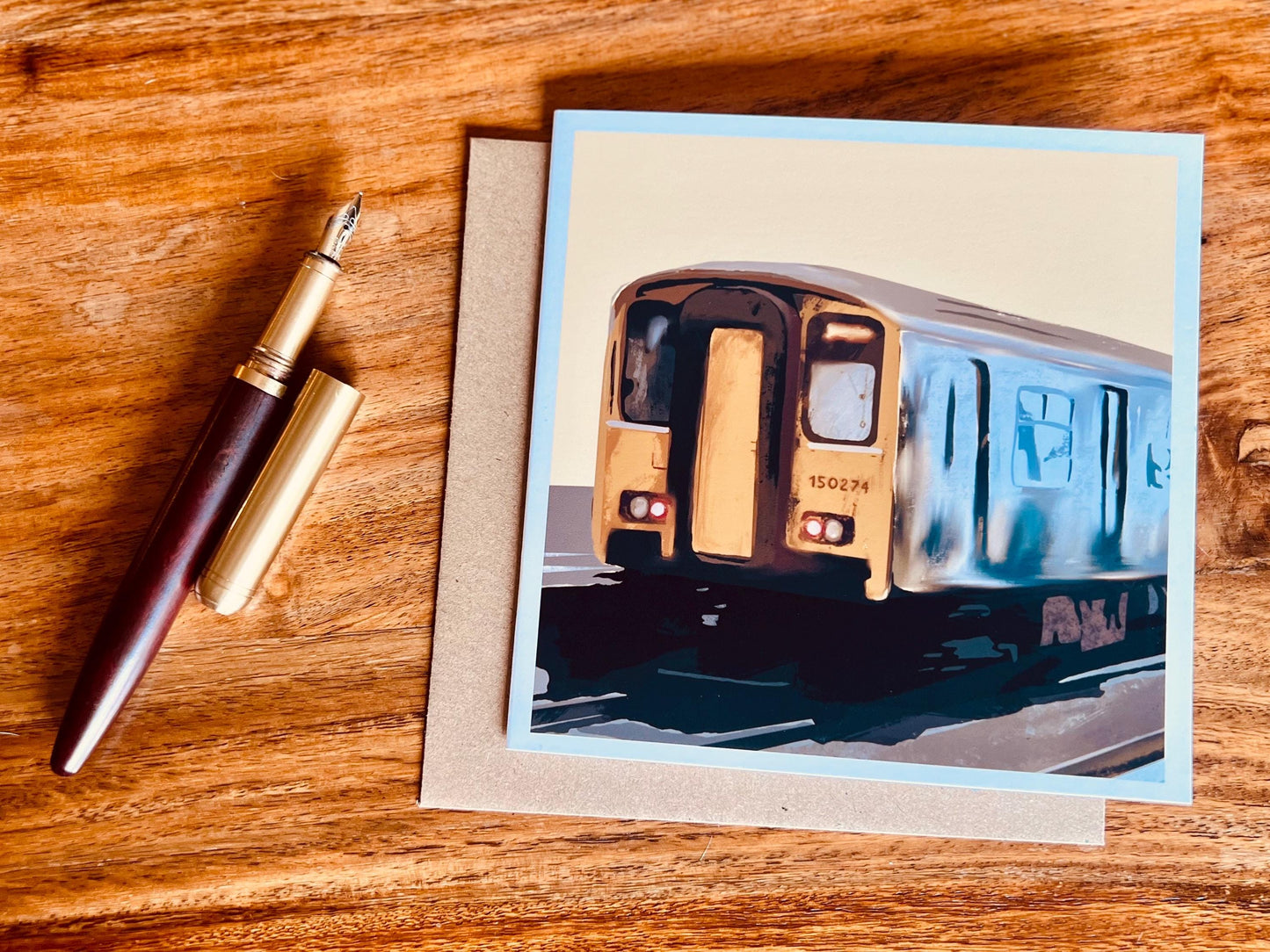 Artisan greeting card featuring Class 150 train 150274 on a yellow background, shown laid flat on top of a kraft envelope on a tabletop next to a pen in a styled flat lay composition.