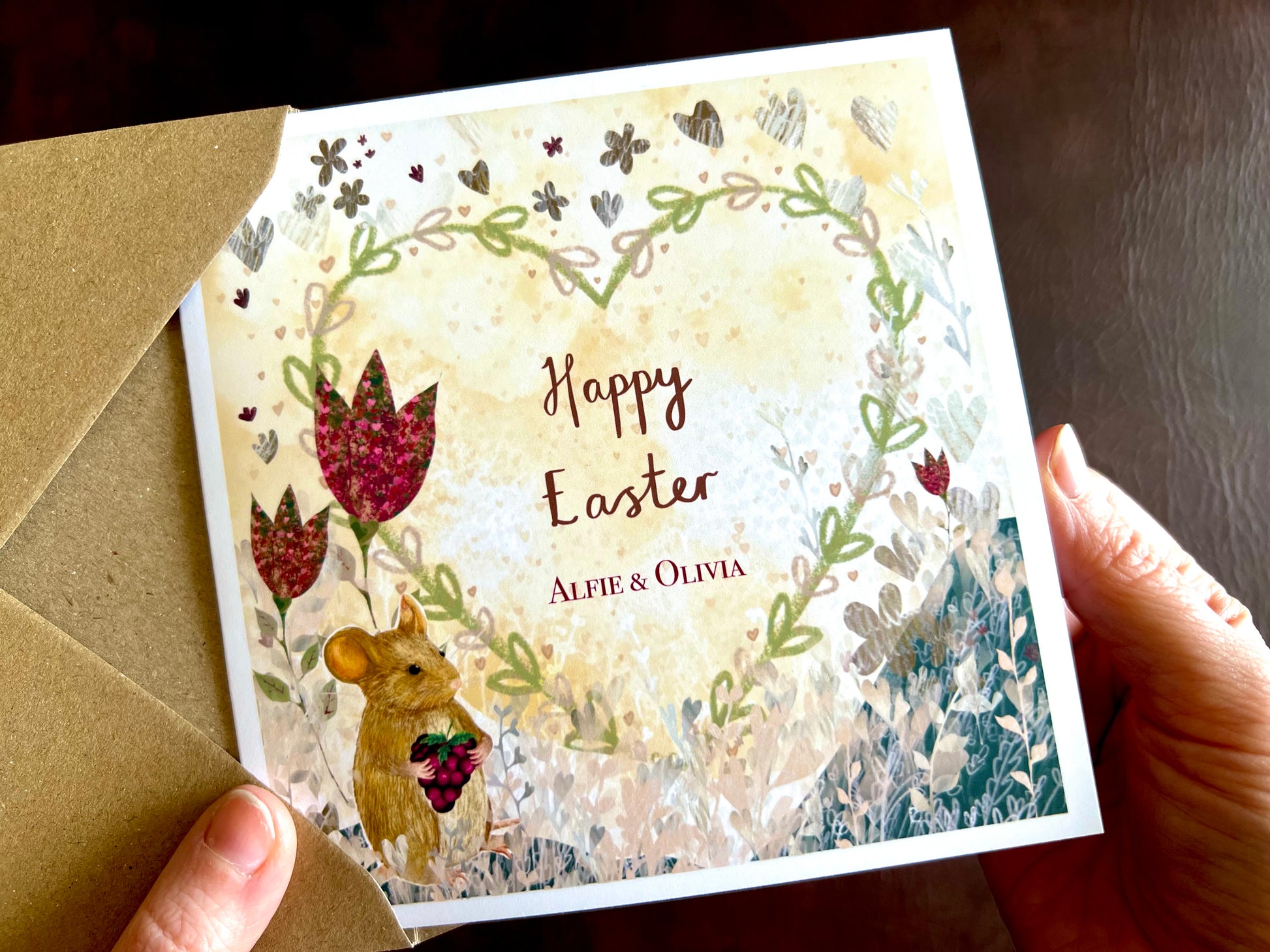 Hands holding an artisan Easter card being removed from a kraft envelope, featuring an illustrated mouse holding a blackberry with red collage style flowers and a floral heart on a lemon yellow background. The card reads 'Happy Easter' followed by two custom names for personalisation.