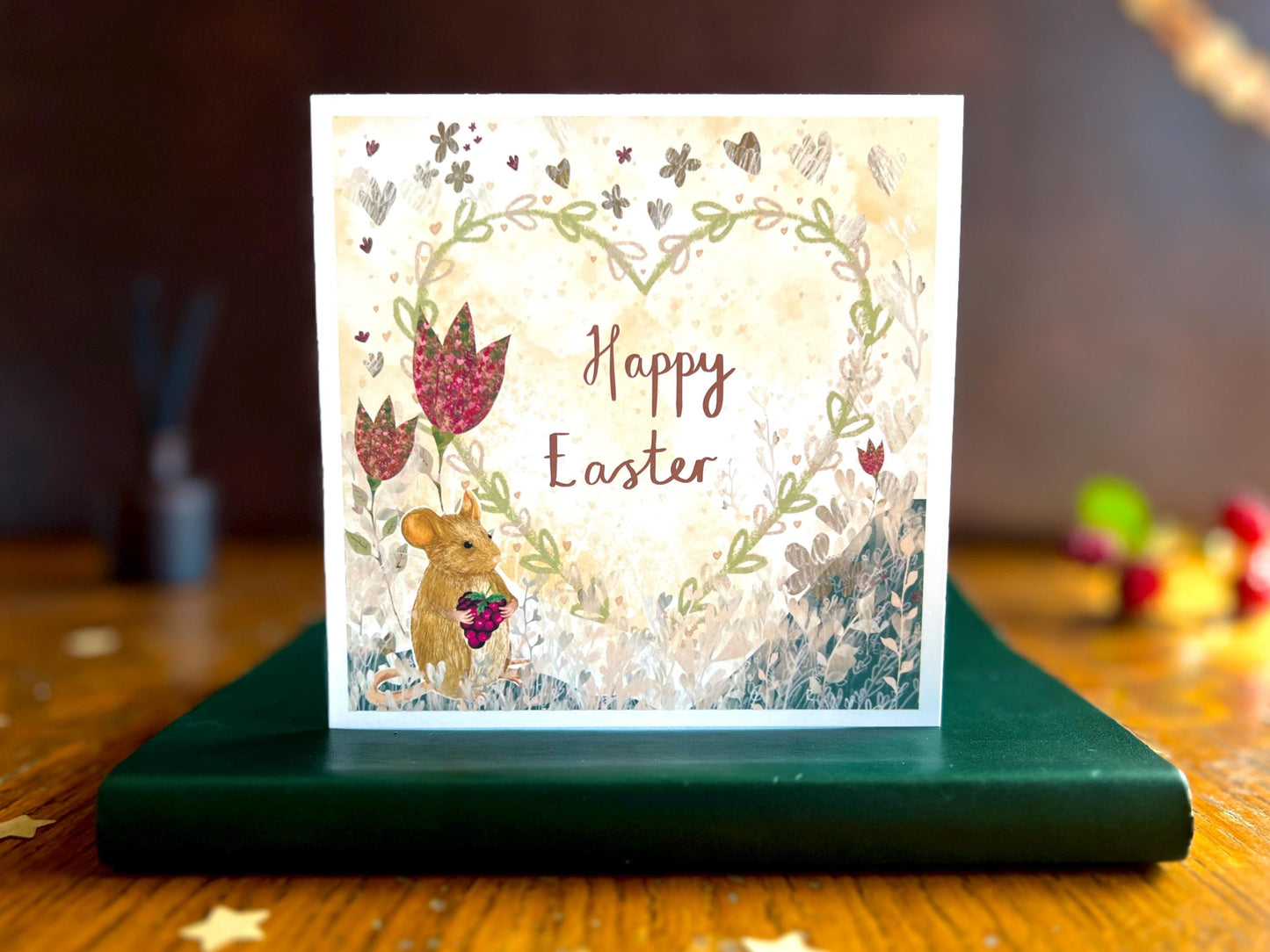Artisan Easter card featuring an illustrated mouse holding a blackberry with red collage style flowers and a floral heart on a lemon yellow background, shown standing upright on a green leatherette book in a styled lifestyle setting. The card reads 'Happy Easter'.
