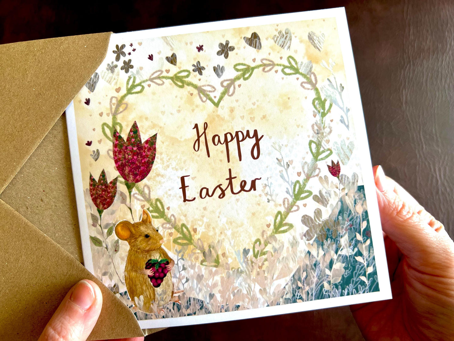 Hands holding an artisan Easter card being removed from a kraft envelope, featuring an illustrated mouse holding a blackberry with red collage style flowers and a floral heart on a lemon yellow background. The card reads 'Happy Easter' in a handwritten style design.