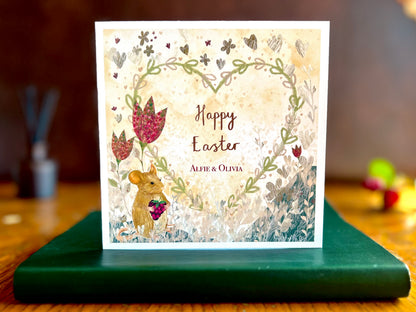 Artisan Easter card featuring an illustrated mouse holding a blackberry with red collage style flowers and a floral heart on a lemon yellow background, shown standing upright on a green leatherette book in a styled lifestyle setting. The design includes custom names for personalisation.