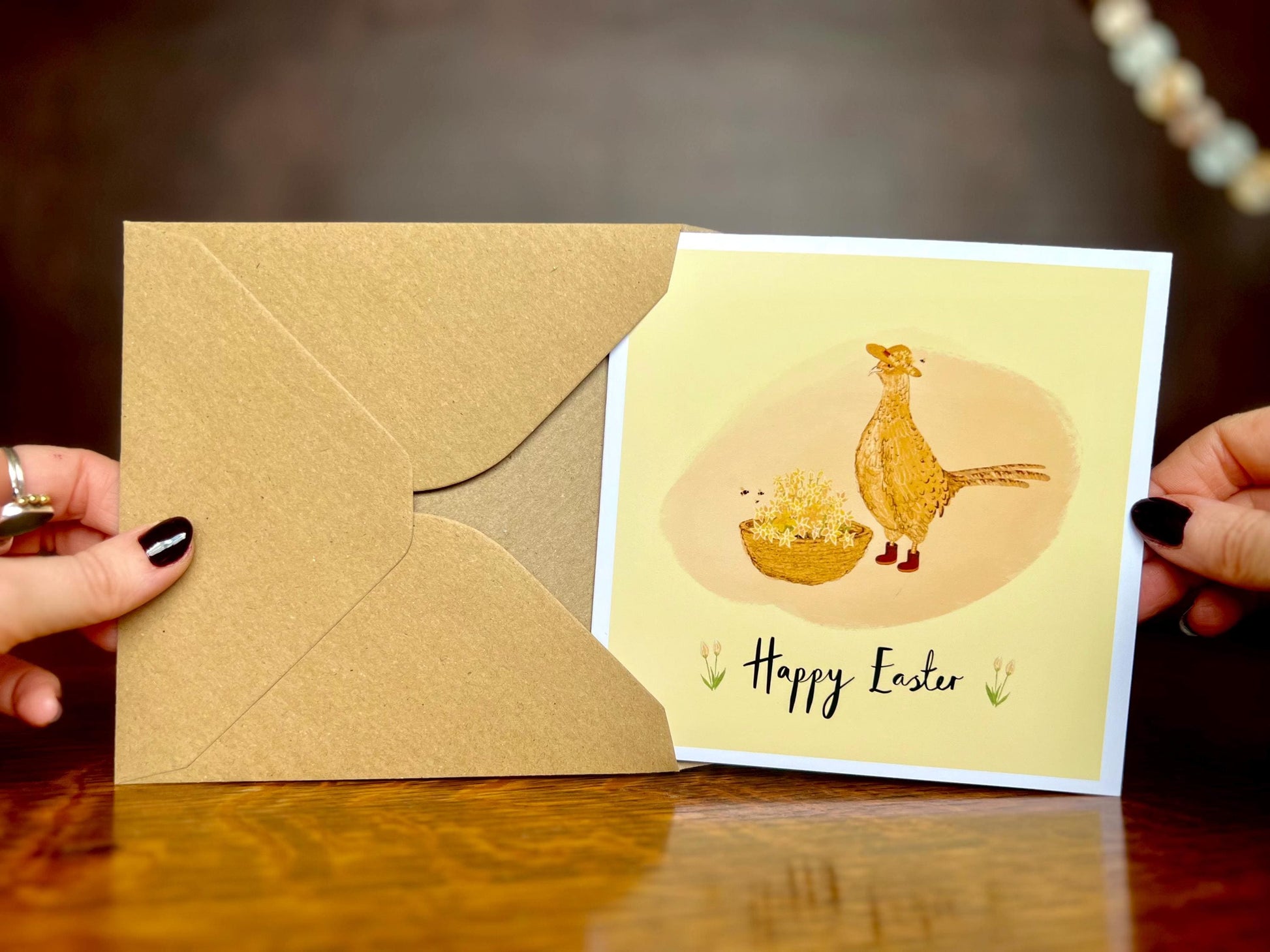 Front view of an artisan Easter card being nearly removed from a kraft envelope, featuring a cheerful pheasant and floral illustration on a yellow background with 'Happy Easter' text shown on the card.