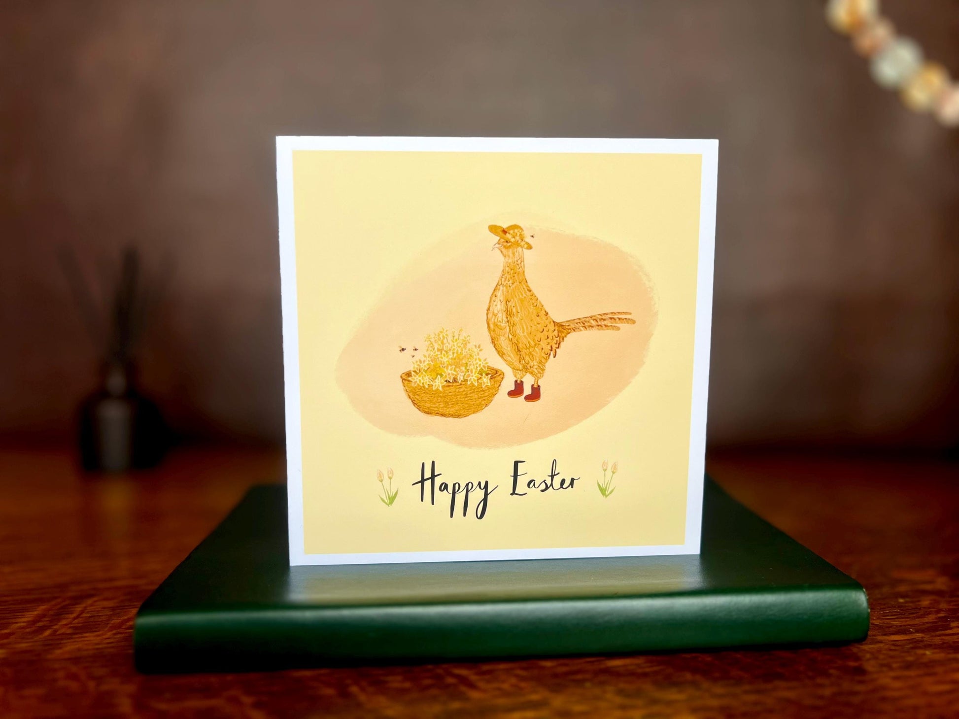 Artisan Easter card featuring a pheasant and floral illustration on a yellow background with 'Happy Easter' text, displayed standing on a green leatherette book on a wooden tabletop with a brown leatherette backdrop.