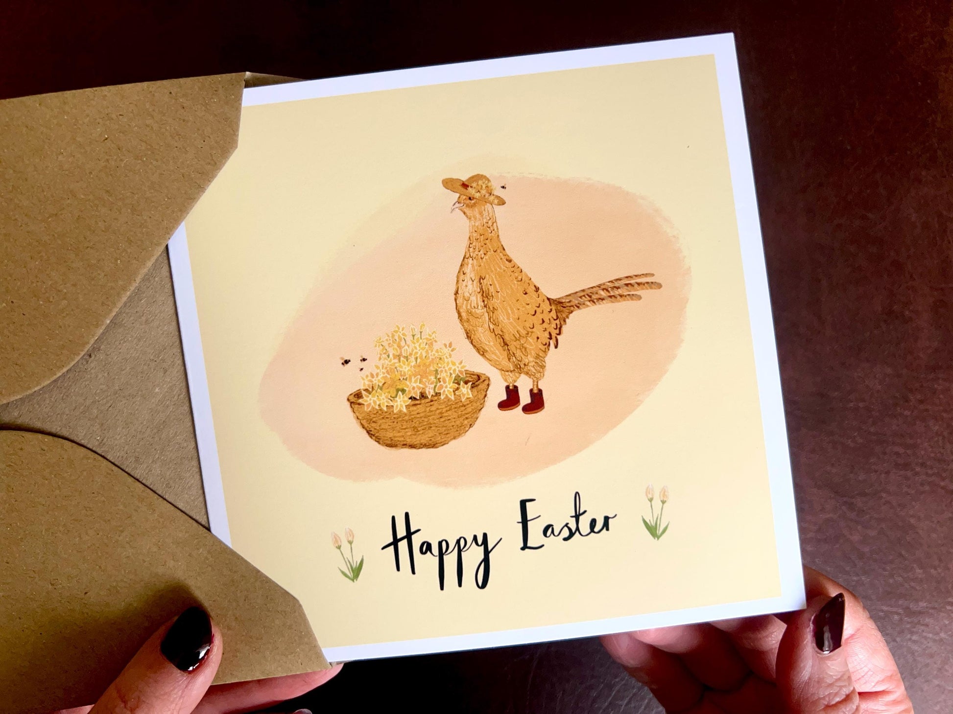 Hands holding an artisan Easter card being removed from a kraft envelope, featuring a cheerful pheasant and floral illustration on a yellow background. The card shows 'Happy Easter' without a custom name, representing the non-personalised version.