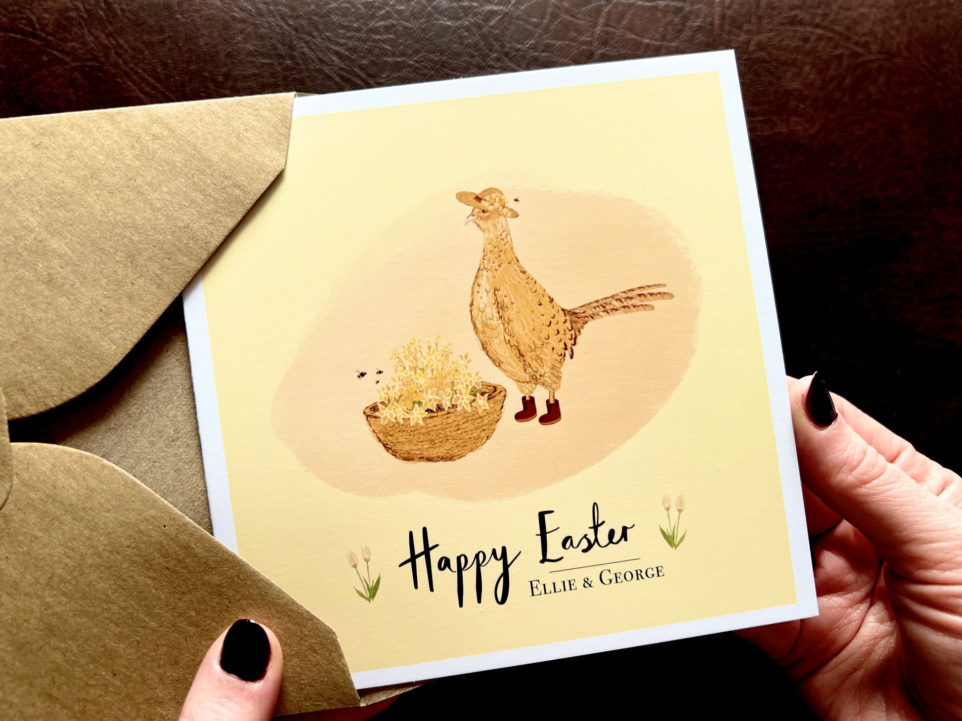Hands holding an artisan Easter card being removed from a kraft envelope, featuring a cheerful pheasant and floral illustration on a yellow background. The card reads 'Happy Easter' with an optional custom name underneath for personalisation.