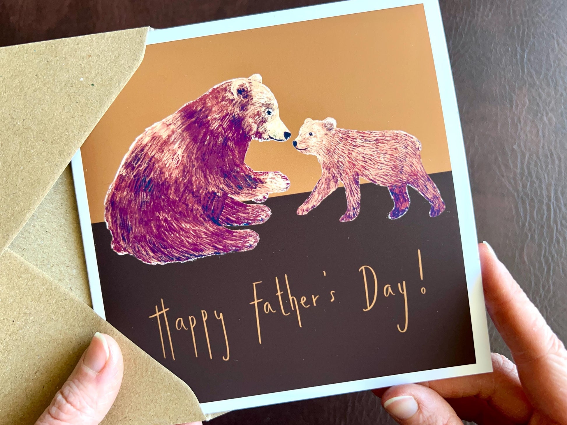 Hands holding an artisan Fathers Day card being removed from a kraft envelope, featuring a brown bear and bear cub illustration on a warm brown and burnt orange background. The card reads 'Happy Father’s Day!' in a celebration design suitable for Father’s Day gifting.