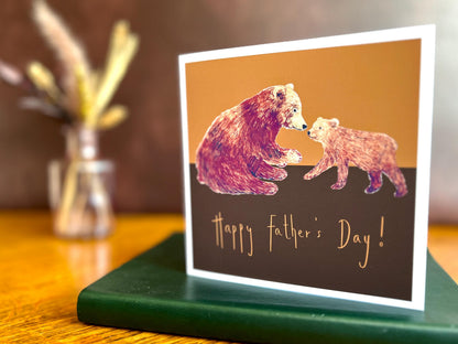 Artisan Fathers Day card featuring a brown bear and bear cub on a warm brown and burnt orange background, shown standing upright on a green leatherette book in a styled lifestyle display.